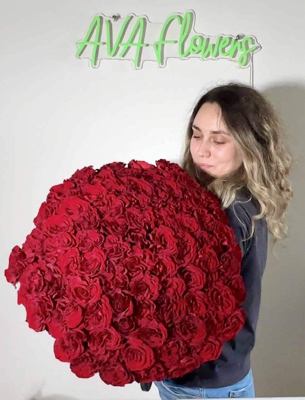 Woman holding a giant bouquet of 101 premium red roses with AVA Flowers sign in the background.