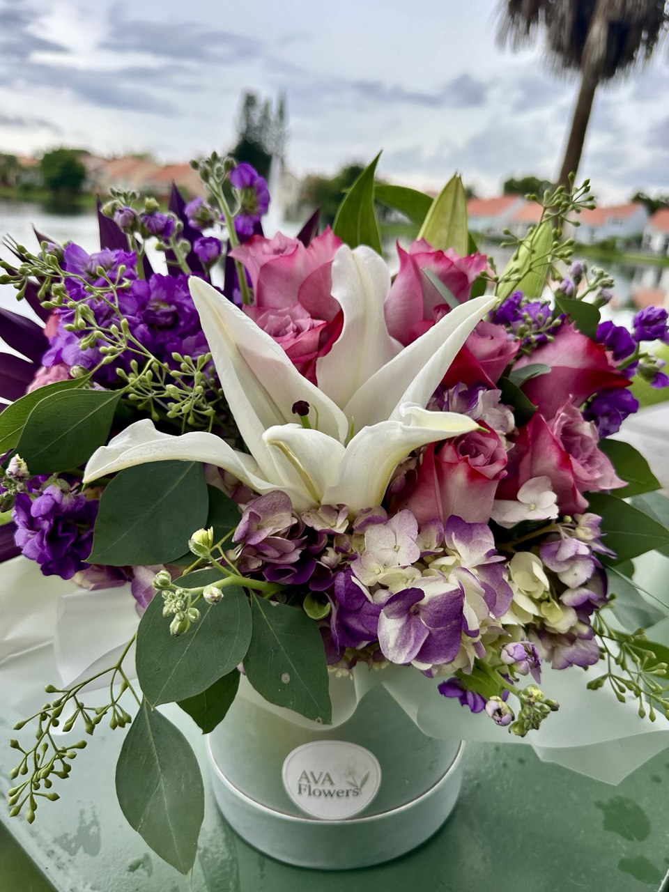 Luxury hat box flower arrangement featuring white lilies, pink roses, hydrangea and vibrant purple seasonal flowers with lush greenery in a silver designer box.
