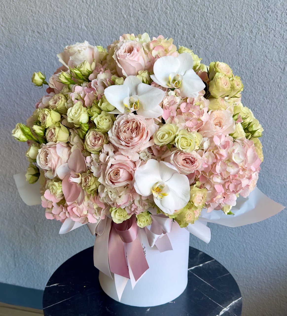 Luxurious pastel flower box with soft pink roses, delicate hydrangeas, spray roses and elegant white orchids, arranged in a round designer hatbox with satin ribbon.