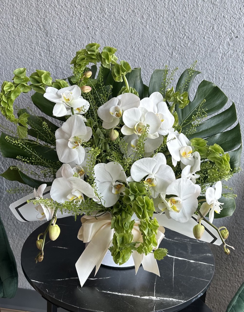 Luxurious hat box arrangement with cascading white Phalaenopsis orchids, lush tropical greenery and decorative leaves, styled with a satin ivory ribbon.