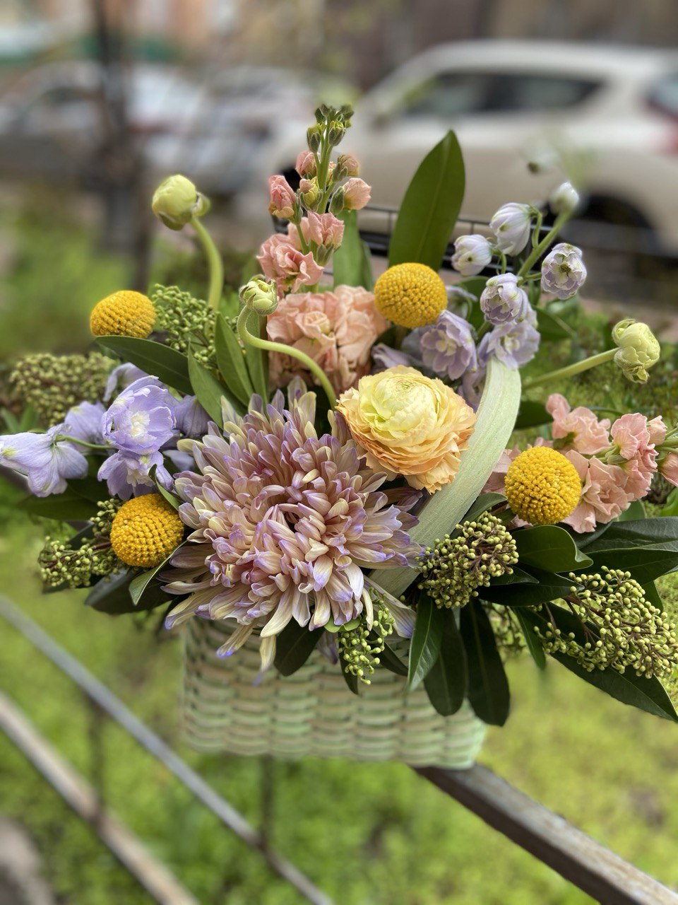 Elegant basket arrangement with pastel chrysanthemums, yellow ranunculus, peach spray roses, purple blooms and round yellow craspedia accents with lush greenery.