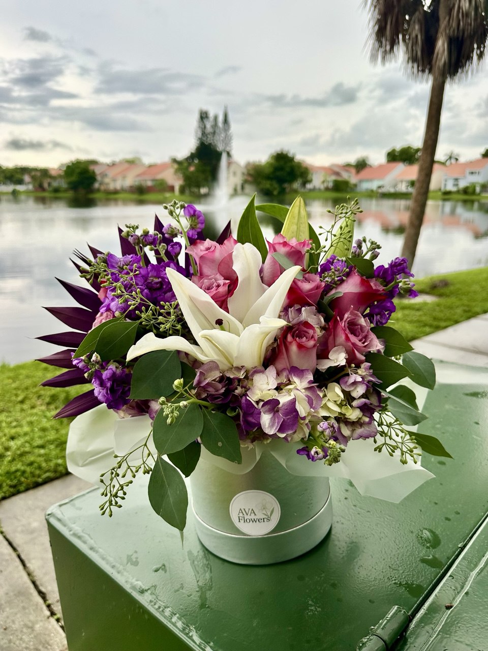 Luxury hat box flower arrangement featuring white lilies, pink roses, hydrangea and vibrant purple seasonal flowers with lush greenery in a silver designer box.