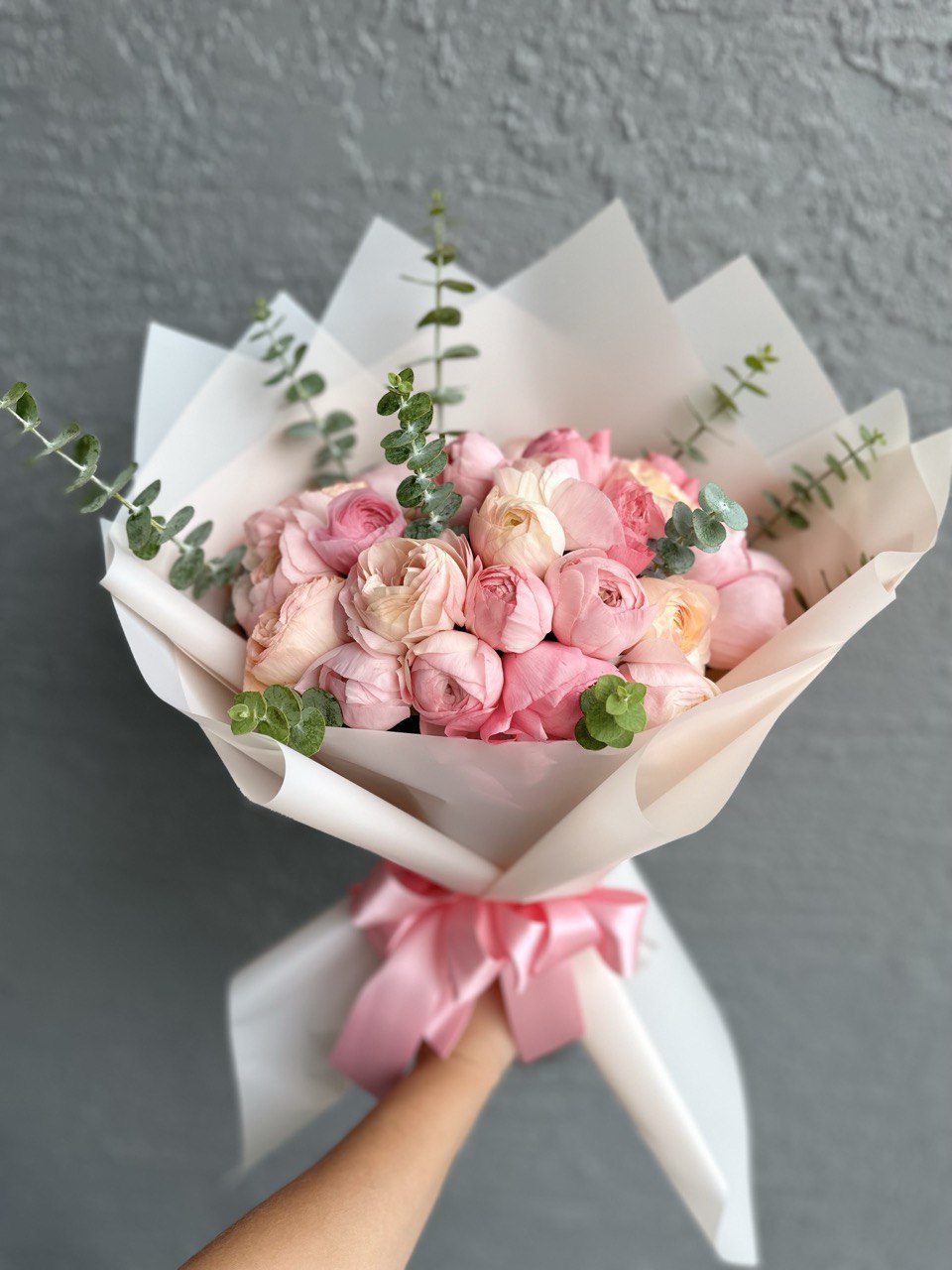 Mono bouquet of pink ranunculus with fresh eucalyptus, wrapped in white paper and tied with a pink satin ribbon — an elegant and romantic floral gift.
