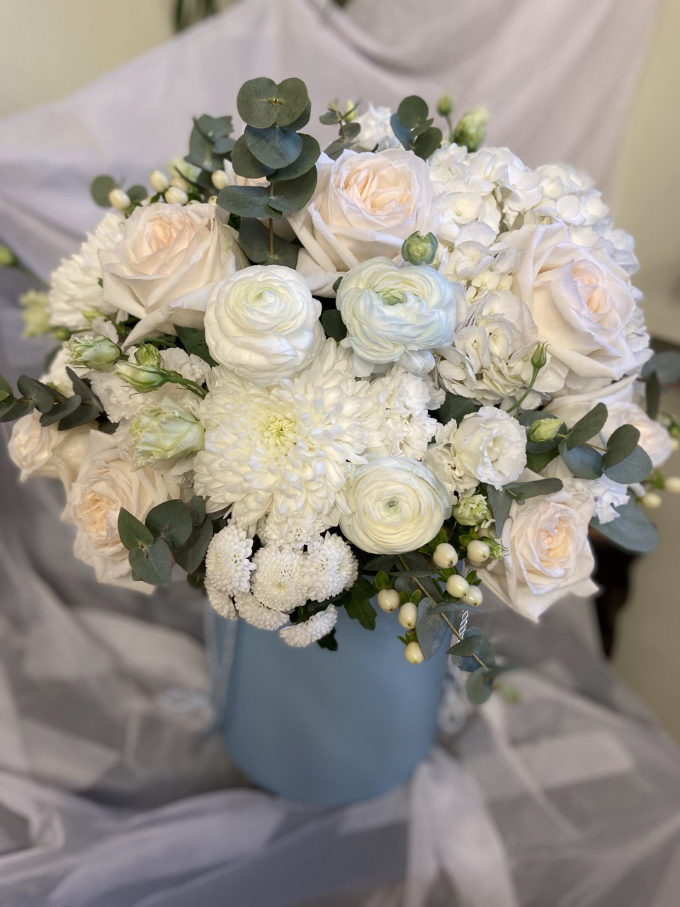 Elegant hatbox flower arrangement with white roses, ranunculus, hydrangeas, chrysanthemums and eucalyptus, creating a fresh and timeless design.