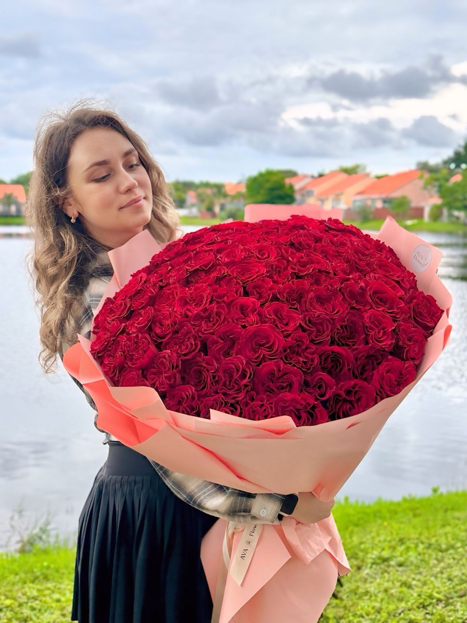 Woman holding a giant bouquet of 101 premium red roses with AVA Flowers sign in the background.