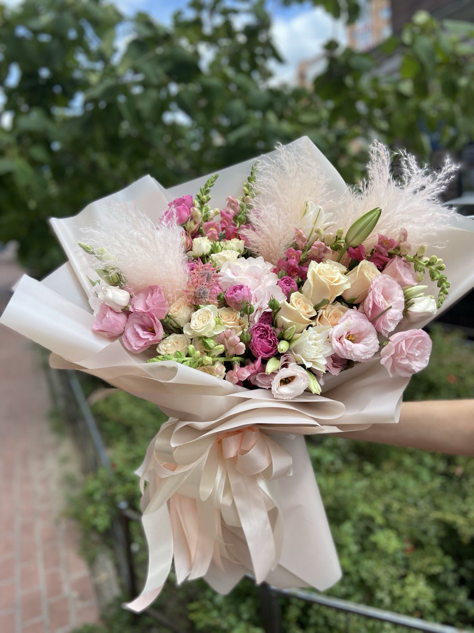 Tender Bliss bouquet with pastel roses, pink lisianthus, spray roses, lilies, and pampas grass wrapped in soft beige paper with satin ribbons — a delicate and romantic floral arrangement