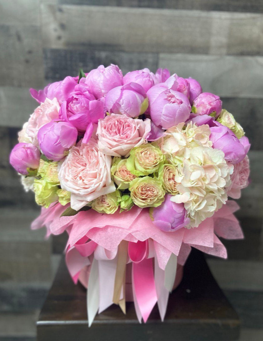 Luxurious hatbox arrangement with lush pink peonies, pastel garden roses, spray roses and hydrangeas, accented with decorative wrapping and satin ribbons.