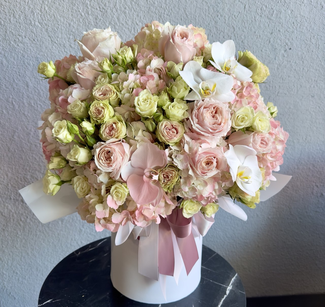 Luxurious pastel flower box with soft pink roses, delicate hydrangeas, spray roses and elegant white orchids, arranged in a round designer hatbox with satin ribbon.