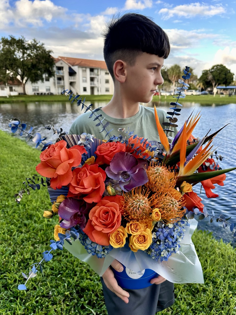 Boy holding the bright "Sunset Glow" flower arrangement with roses, orchids, protea, and bird of paradise in a blue hatbox by AvaFlowers near the lake