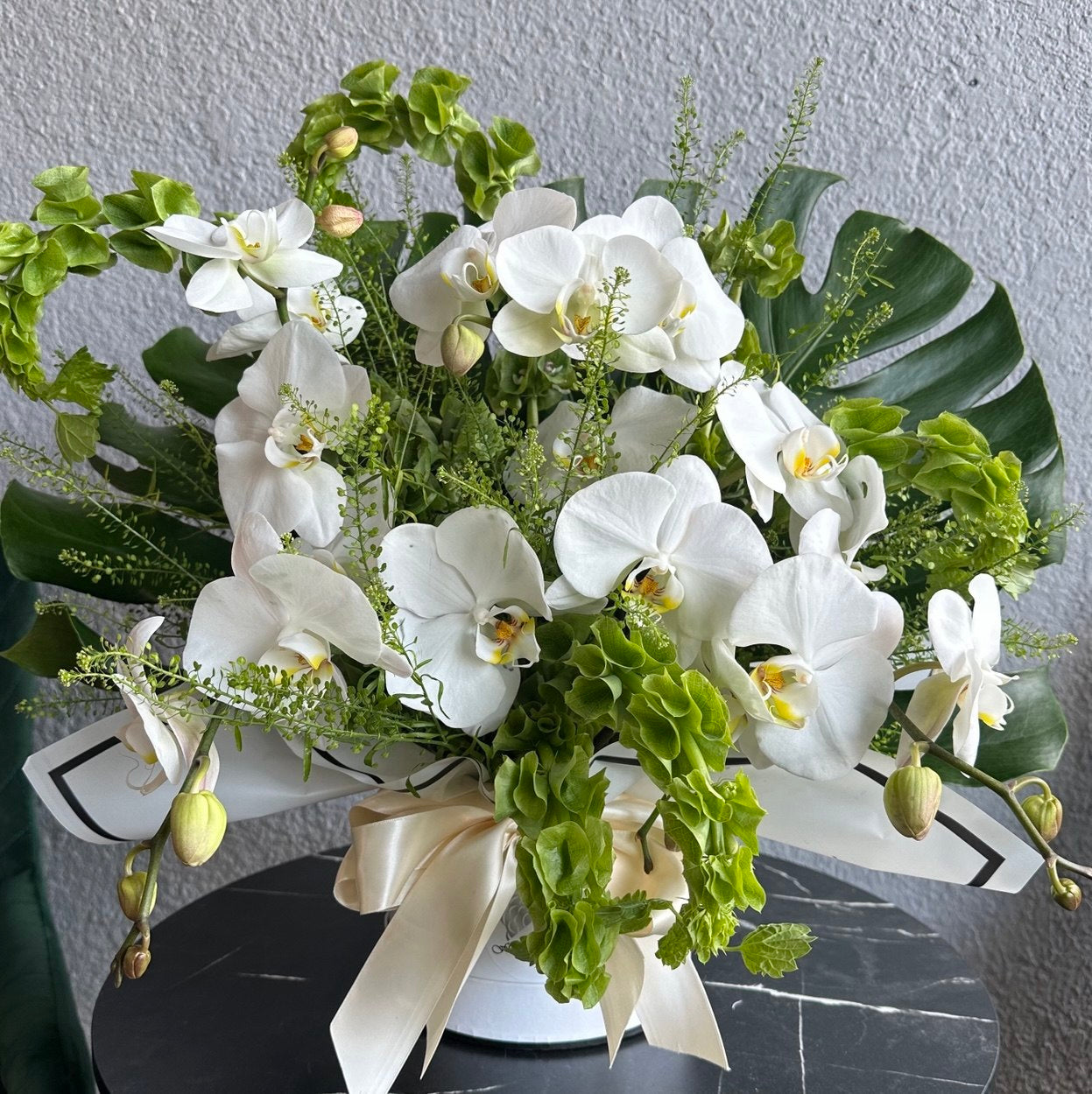 Luxurious hat box arrangement with cascading white Phalaenopsis orchids, lush tropical greenery and decorative leaves, styled with a satin ivory ribbon.