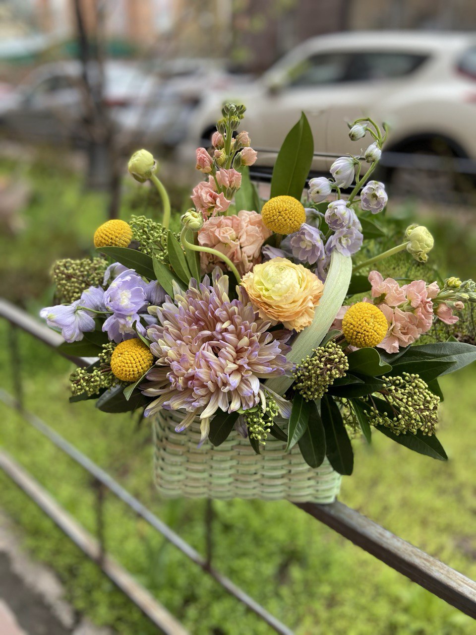 Elegant basket arrangement with pastel chrysanthemums, yellow ranunculus, peach spray roses, purple blooms and round yellow craspedia accents with lush greenery.