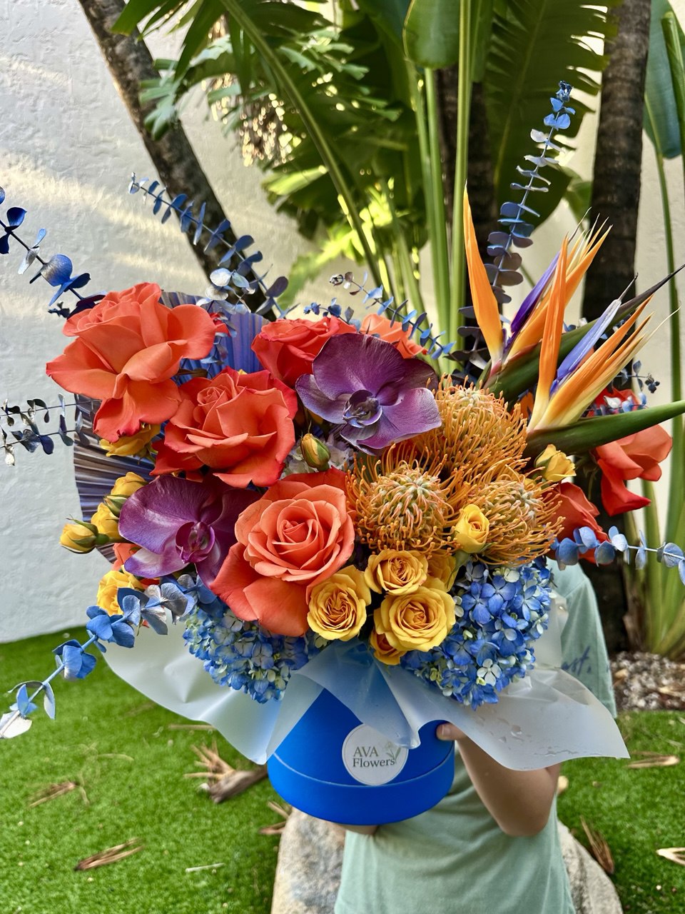 Close-up of the "Sunset Glow" bouquet: roses, orchids, protea, and hydrangea in a blue AvaFlowers hatbox, decorated with eucalyptus