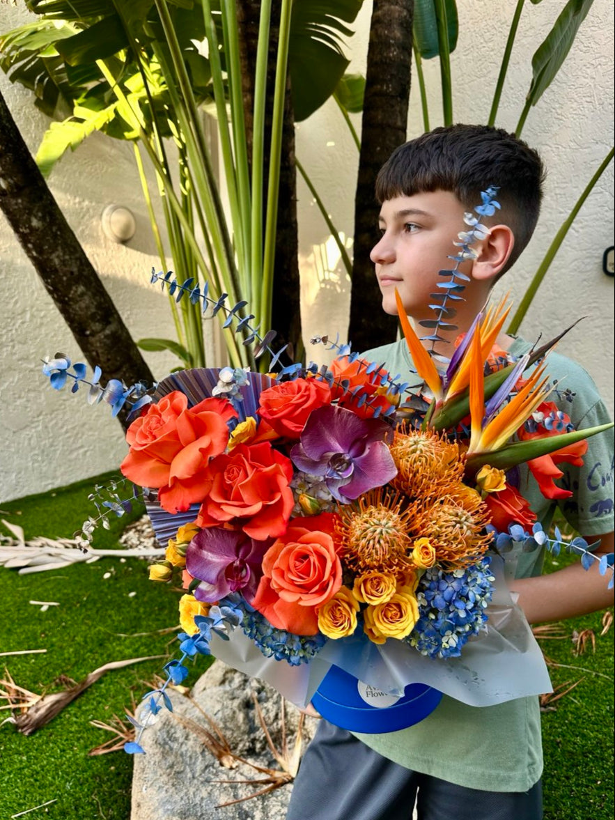 Boy posing with the "Sunset Glow" hatbox flower arrangement, vibrant flowers contrasting with the green lawn and palm trees in the background.