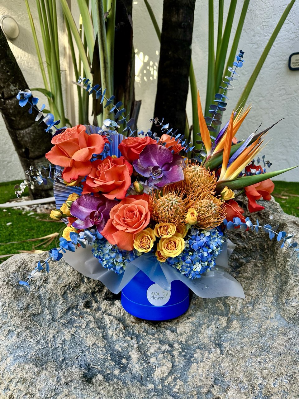 Close-up of the "Sunset Glow" bouquet: roses, orchids, protea, and hydrangea in a blue AvaFlowers hatbox, decorated with eucalyptus