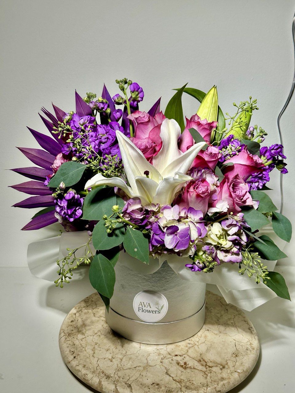 Luxury hat box flower arrangement featuring white lilies, pink roses, hydrangea and vibrant purple seasonal flowers with lush greenery in a silver designer box.