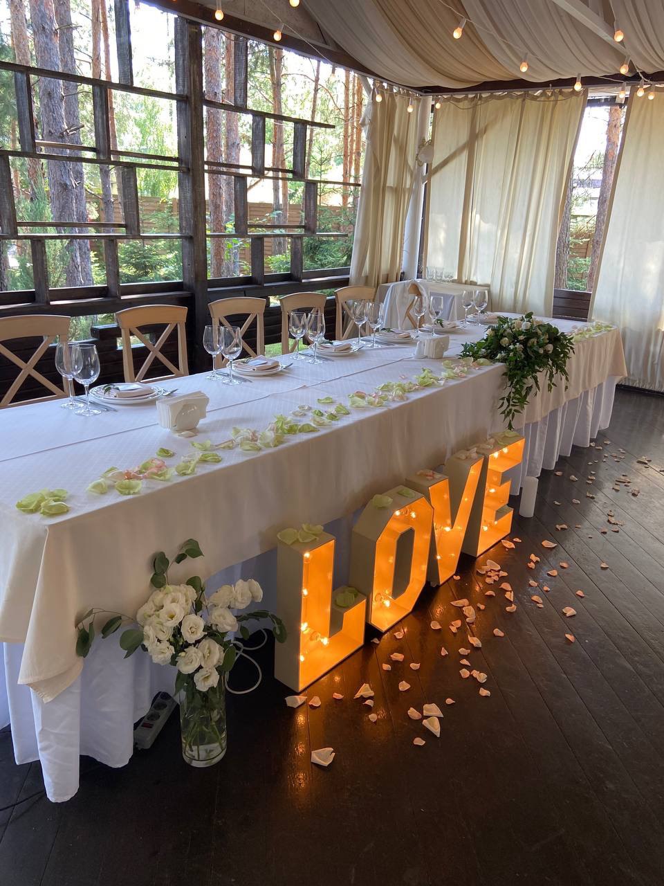 Elegant wedding head table decorated with fresh flowers, glowing LOVE marquee letters, rose petals, and greenery for a romantic reception setup.
