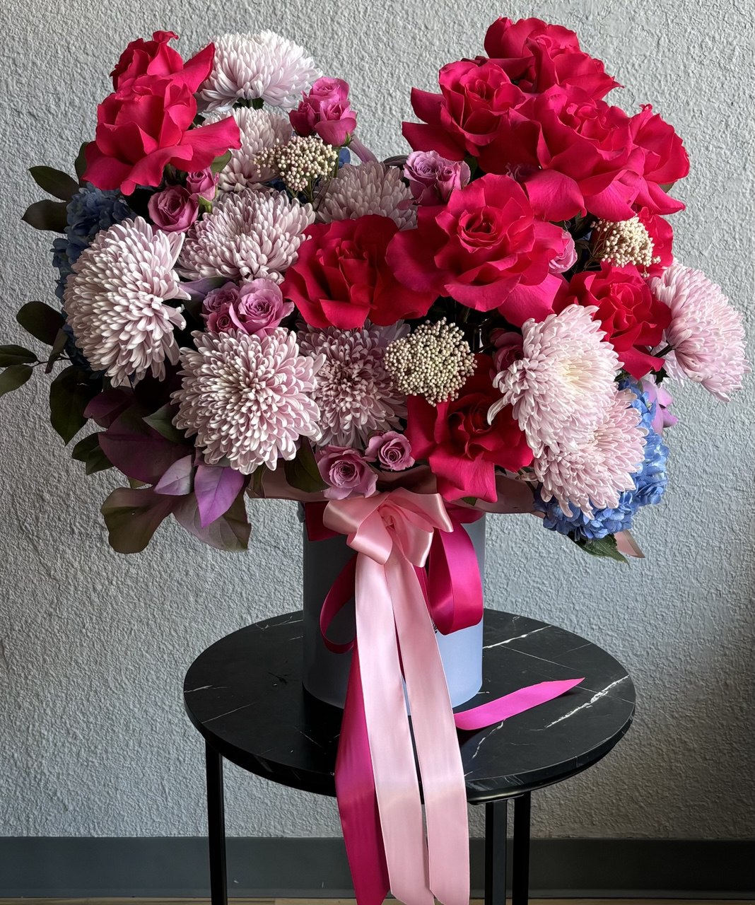 Luxurious flower arrangement in a designer hatbox with bright hot pink roses, pastel chrysanthemums, spray roses and hydrangea, tied with a pink satin ribbon.