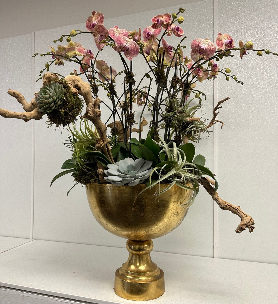 Artistic arrangement of pink orchids, succulents, and air plants with driftwood in a golden vase.