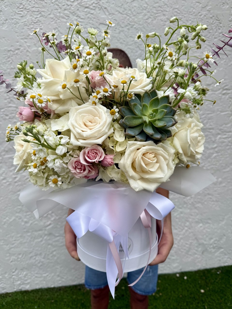 Bouquet of flowers with white roses, pink roses, and succulents held by a person against a plain background.