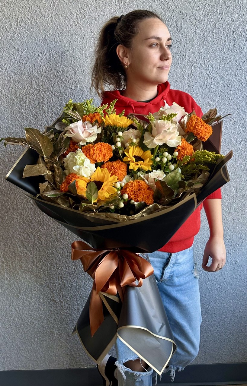 Large autumn-inspired bouquet with bright orange marigolds, yellow sunflowers, cream roses and greenery, wrapped in elegant black paper with a bronze satin ribbon.