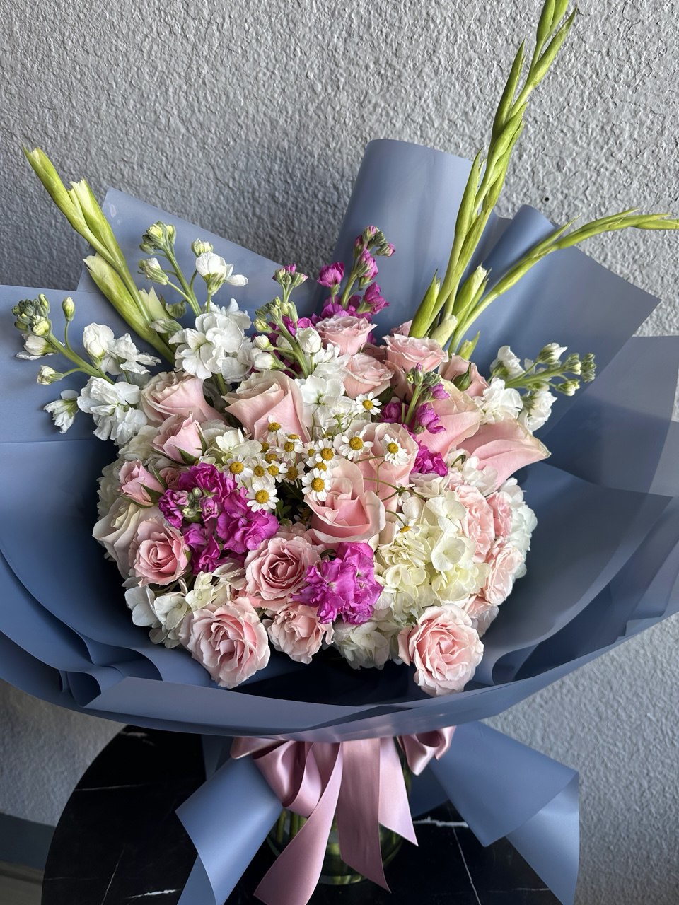 Elegant bouquet with pink roses, white hydrangeas, bright magenta accents and daisies, wrapped in blue paper with a pink ribbon.