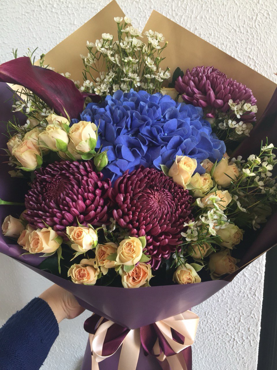 A stylish bouquet featuring deep blue hydrangea, burgundy chrysanthemums, creamy spray roses and white filler flowers, wrapped in purple paper with a satin bow.