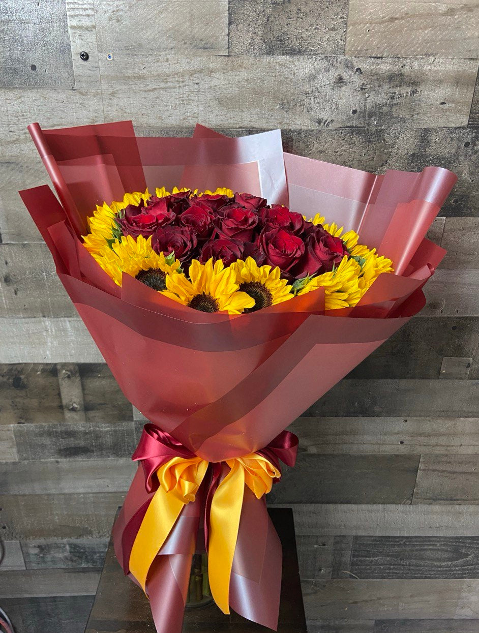 Vibrant bouquet with deep red roses surrounded by bright yellow sunflowers, wrapped in burgundy paper with matching ribbons.