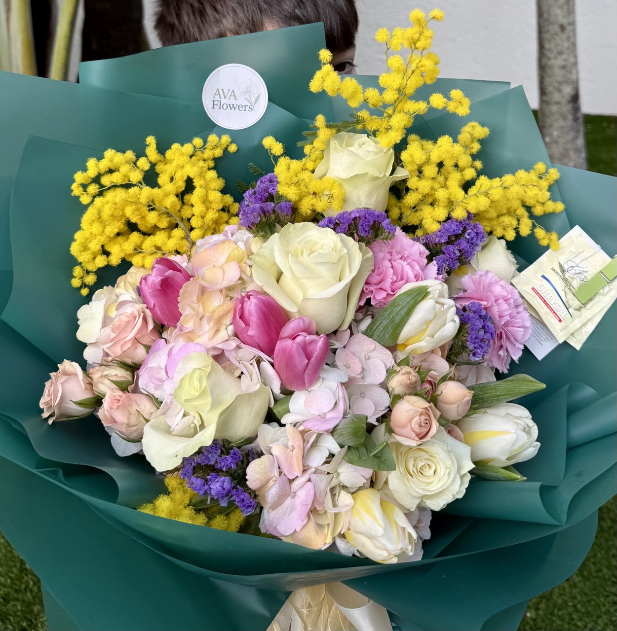 Bouquet of flowers with yellow and purple accents, wrapped in green paper, held by a person.