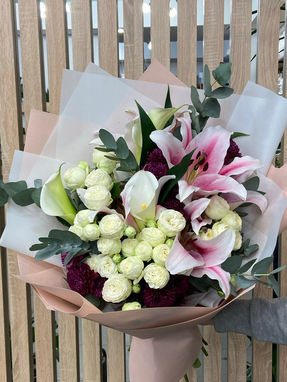 Lily’s Embrace bouquet with elegant pink lilies, delicate white spray roses, calla lilies, and lush eucalyptus, beautifully wrapped in pastel paper for a romantic and graceful floral arrangement