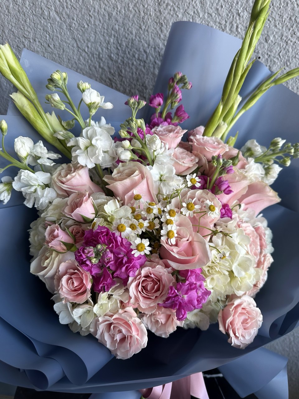 Elegant bouquet with pink roses, white hydrangeas, bright magenta accents and daisies, wrapped in blue paper with a pink ribbon.