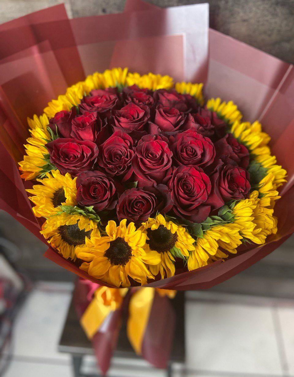 Vibrant bouquet with deep red roses surrounded by bright yellow sunflowers, wrapped in burgundy paper with matching ribbons.