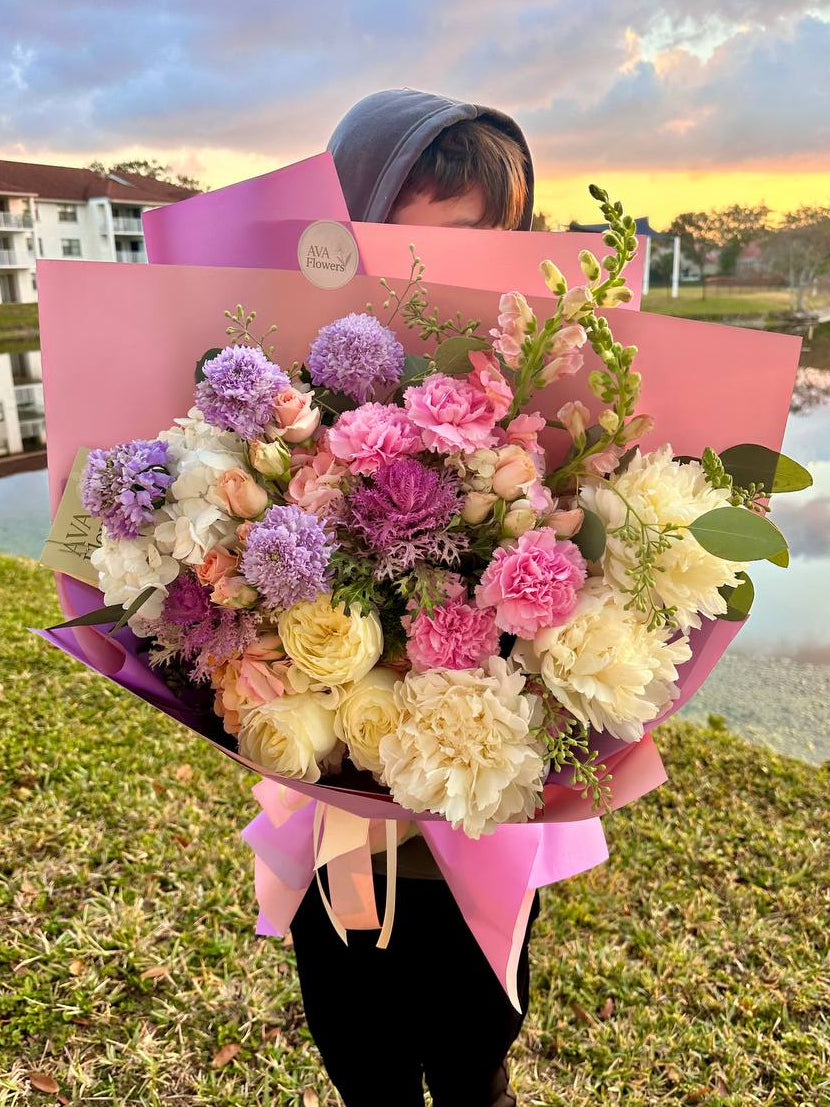 Person holding a large bouquet of flowers with a pink box against a scenic background.