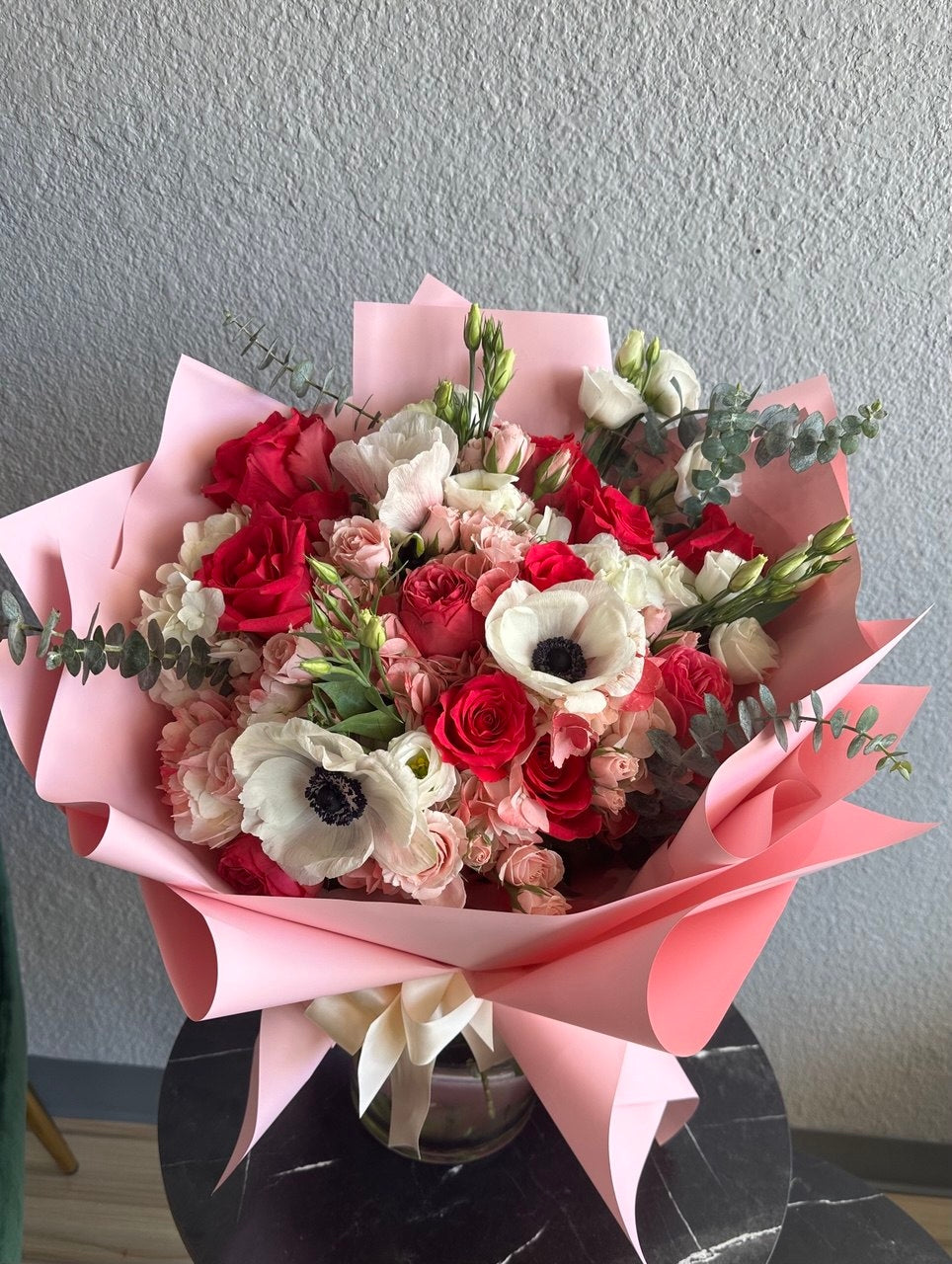 Charming bouquet with pink and red roses, delicate white anemones, spray roses and eucalyptus, wrapped in pastel pink paper and tied with a satin ribbon.