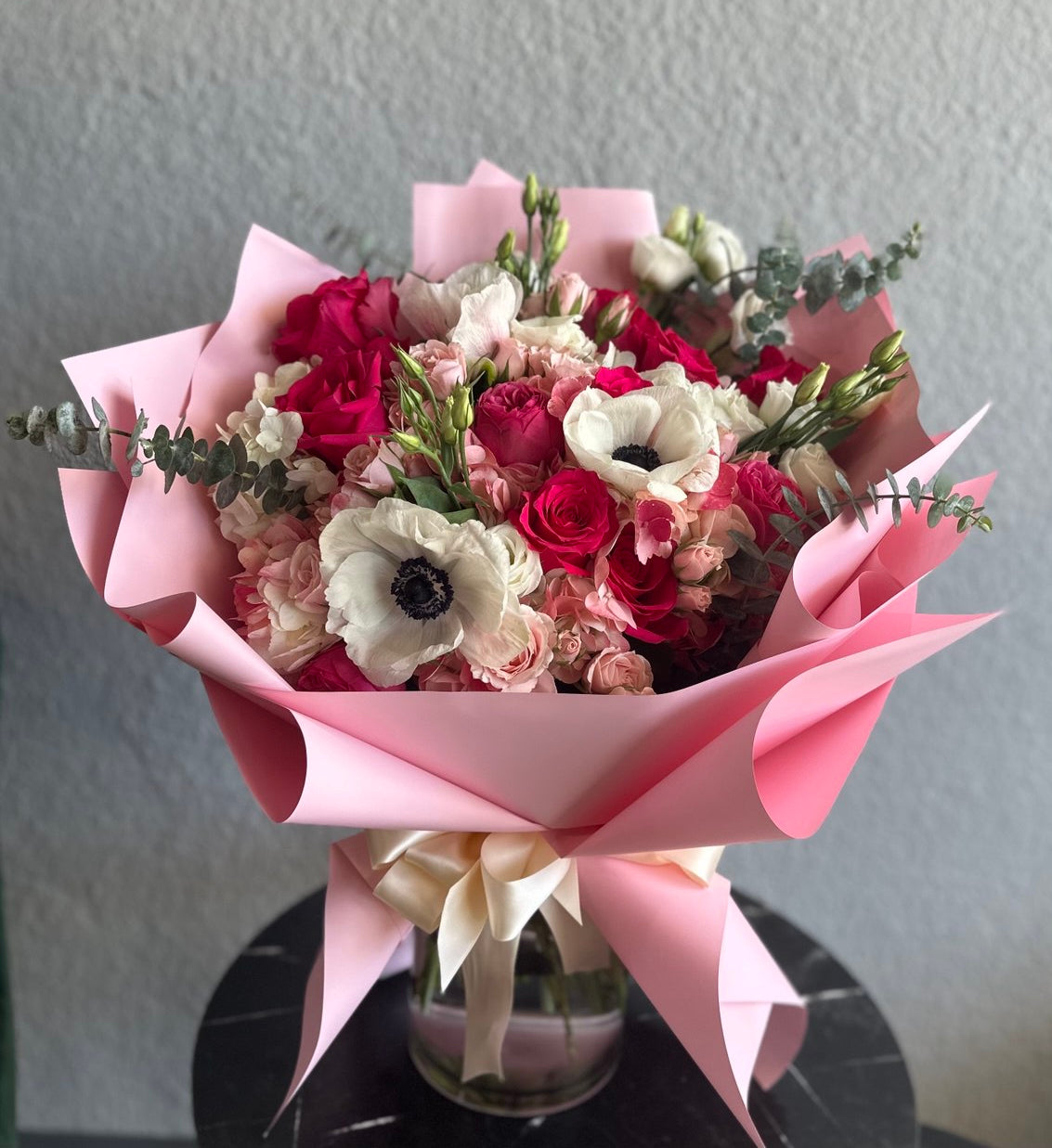 Charming bouquet with pink and red roses, delicate white anemones, spray roses and eucalyptus, wrapped in pastel pink paper and tied with a satin ribbon.