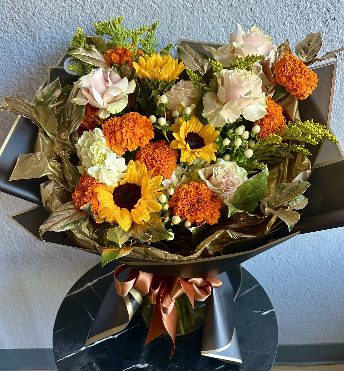 Large autumn-inspired bouquet with bright orange marigolds, yellow sunflowers, cream roses and greenery, wrapped in elegant black paper with a bronze satin ribbon.