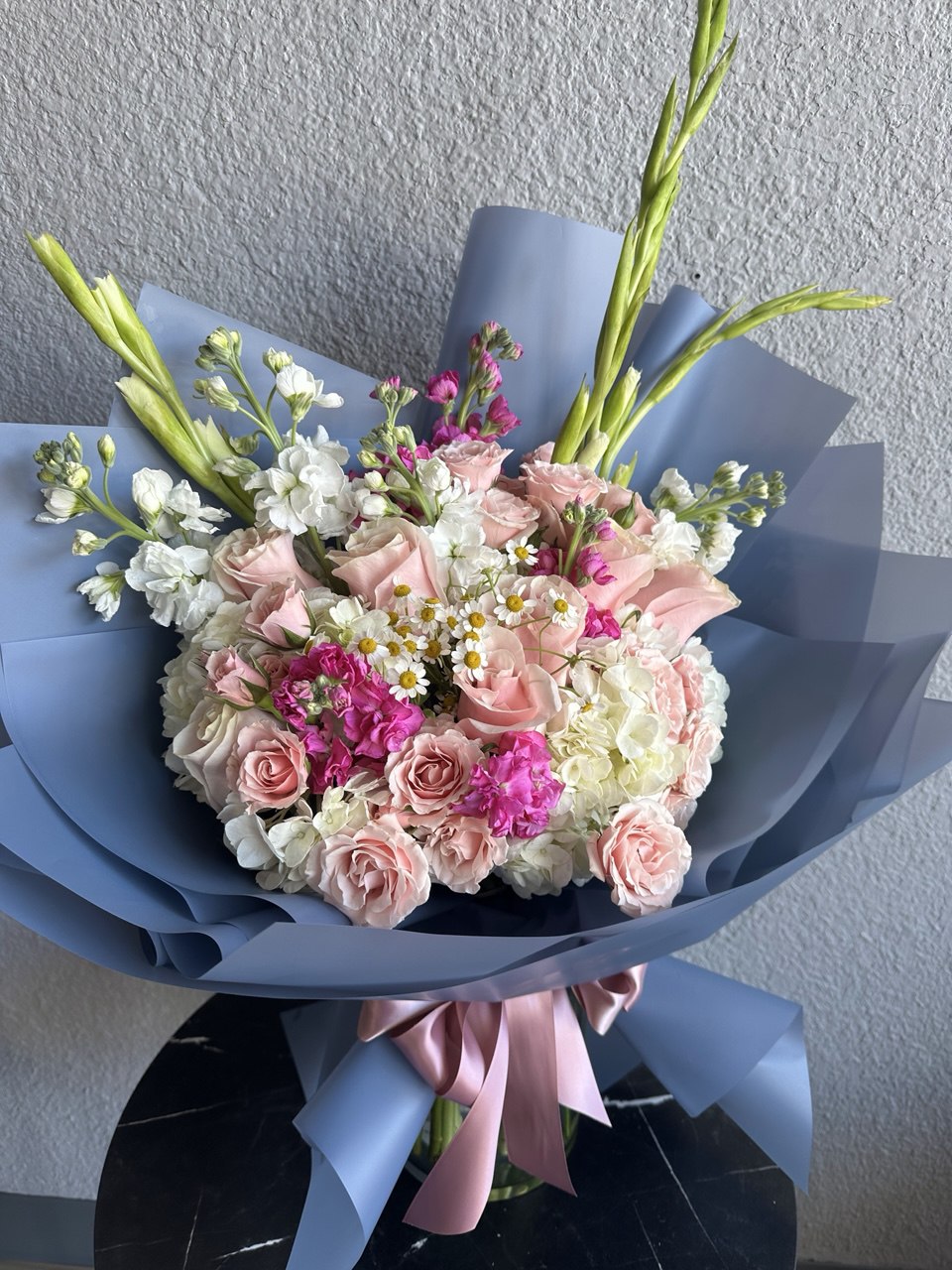 Elegant bouquet with pink roses, white hydrangeas, bright magenta accents and daisies, wrapped in blue paper with a pink ribbon.