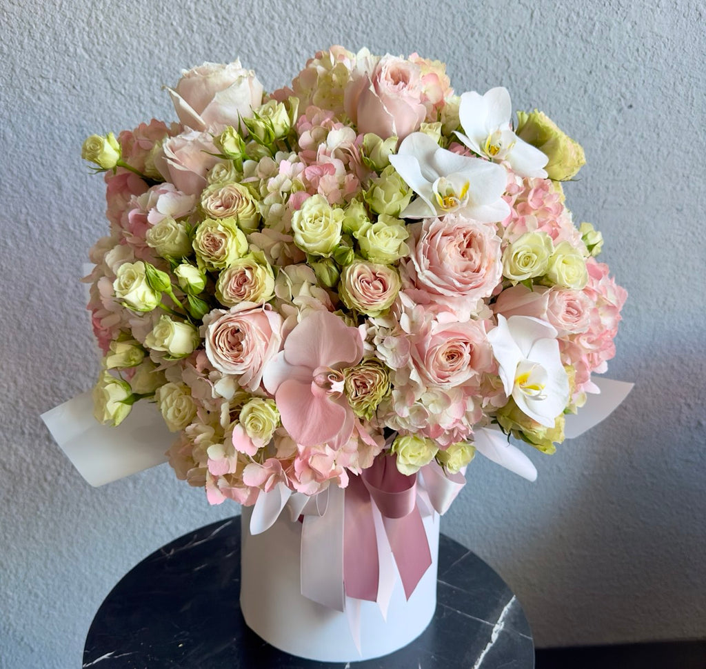 Luxurious pastel flower box with soft pink roses, delicate hydrangeas, spray roses and elegant white orchids, arranged in a round designer hatbox with satin ribbon.