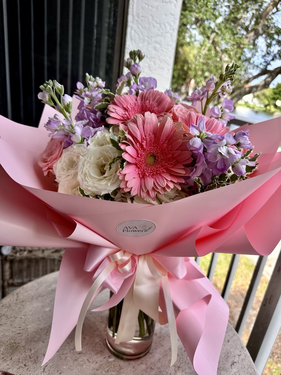 Bouquet of flowers wrapped in pink paper with a visible brand label, set against an outdoor background.