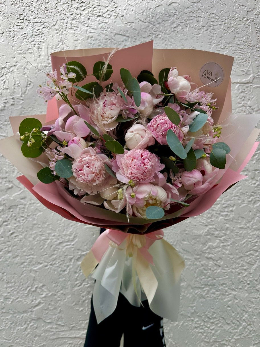 Bouquet of pink flowers wrapped in brown paper with a white ribbon against a white wall.