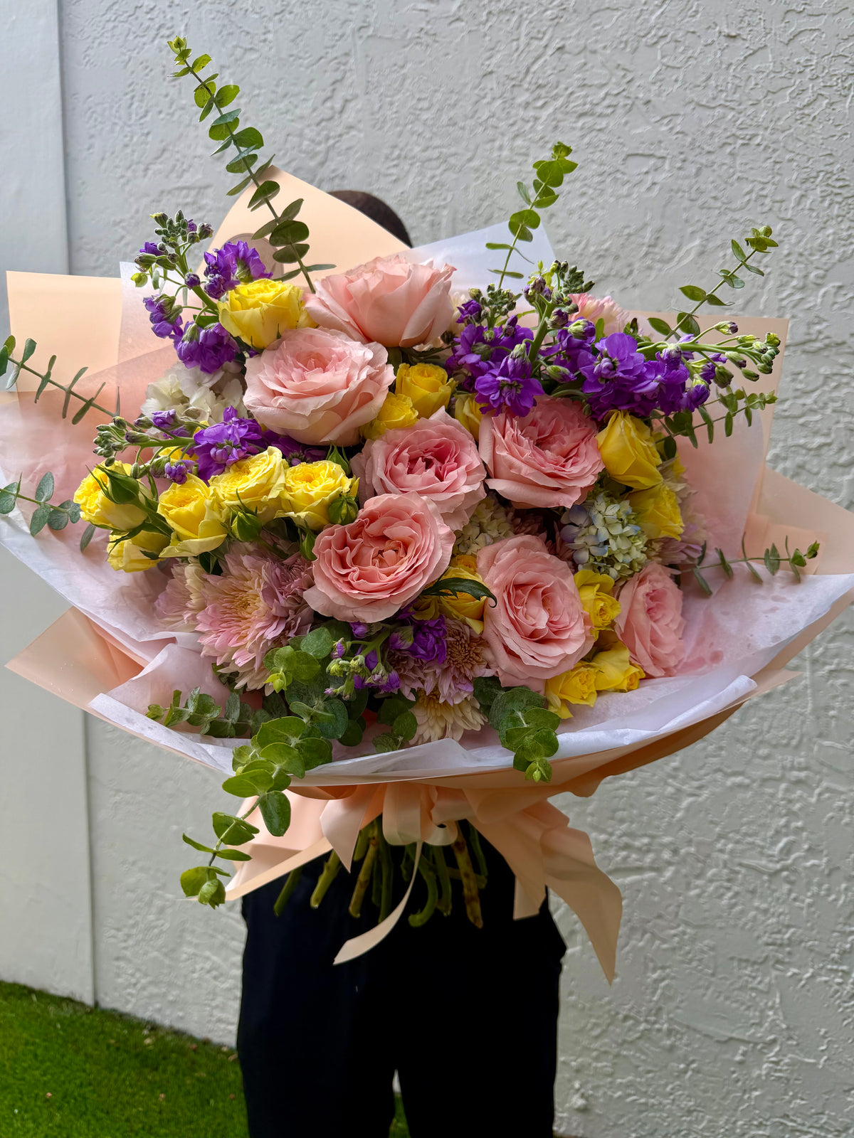 A large, vibrant floral bouquet held by a person against a textured white wall. The bouquet features pink roses, yellow roses, purple flowers, and green foliage, wrapped in peach-colored paper with a peach ribbon.