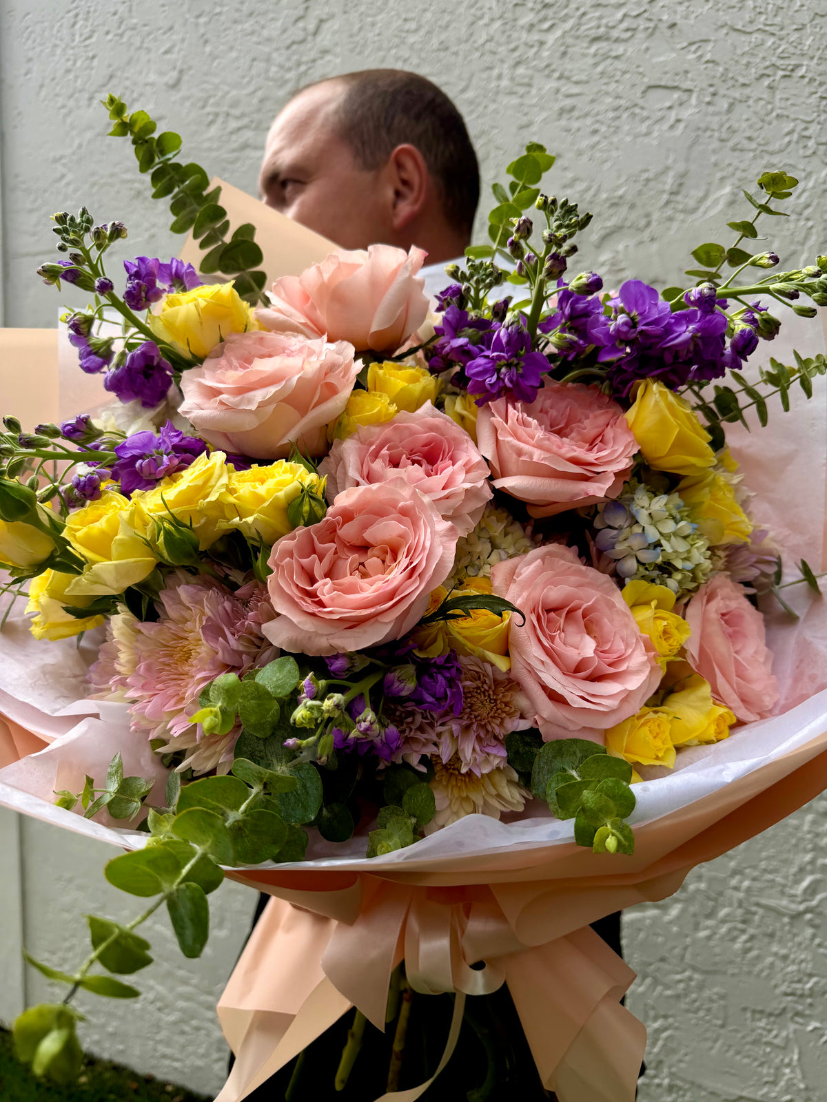 A large, vibrant flower bouquet featuring pink roses, yellow carnations, purple stock, and green foliage, wrapped in white tissue paper with a peach ribbon. A man is partially visible in the background holding a box against a textured light gray wall.