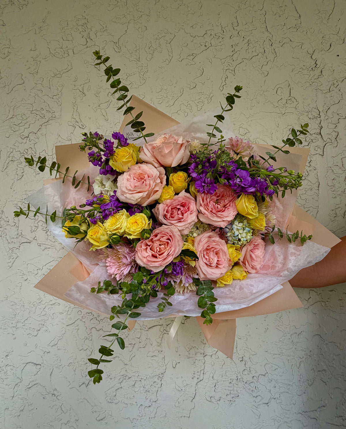 A vibrant floral bouquet featuring pink roses, yellow roses, purple flowers, and green eucalyptus stems, wrapped in brown kraft paper and clear cellophane, held against a textured off-white wall.