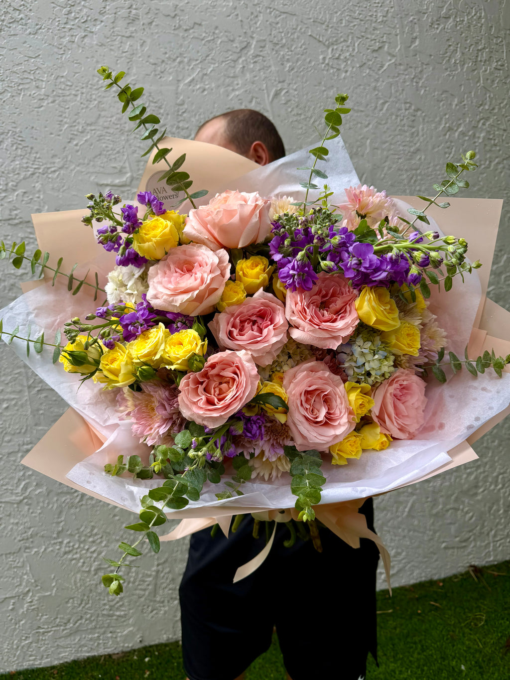 A large, vibrant flower bouquet held by a person whose head is hidden behind it. The bouquet features pink roses, yellow carnations, purple stock, and green eucalyptus stems, wrapped in white and beige paper.