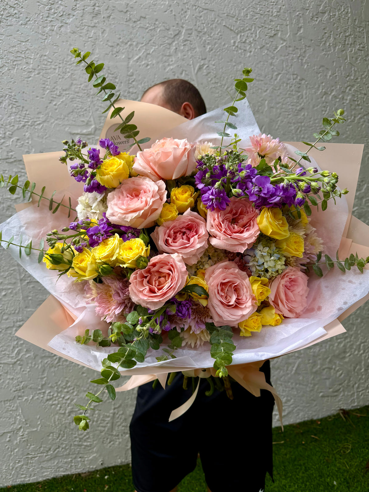 A large, vibrant flower bouquet held by a person whose head is hidden behind it. The bouquet features pink roses, yellow carnations, purple stock, and green eucalyptus stems, wrapped in white and beige paper.