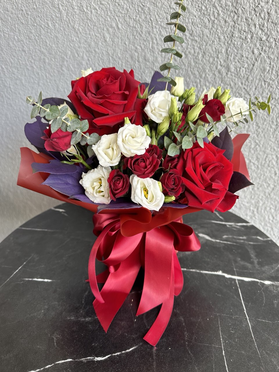 Elegant hand-tied bouquet with deep red roses, spray roses, white lisianthus and eucalyptus, wrapped in red and purple designer paper with a satin ribbon bow