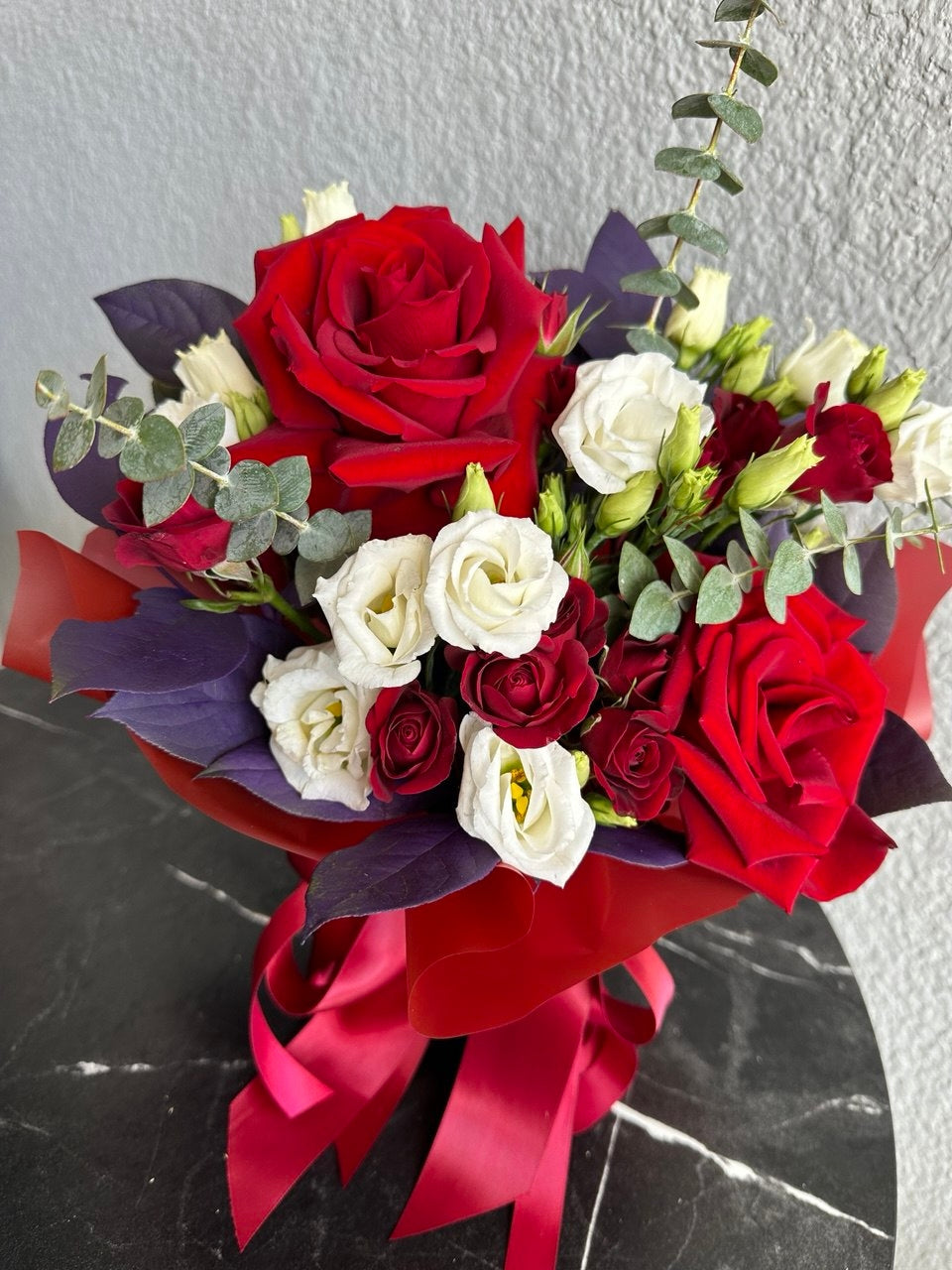 Elegant hand-tied bouquet with deep red roses, spray roses, white lisianthus and eucalyptus, wrapped in red and purple designer paper with a satin ribbon bow