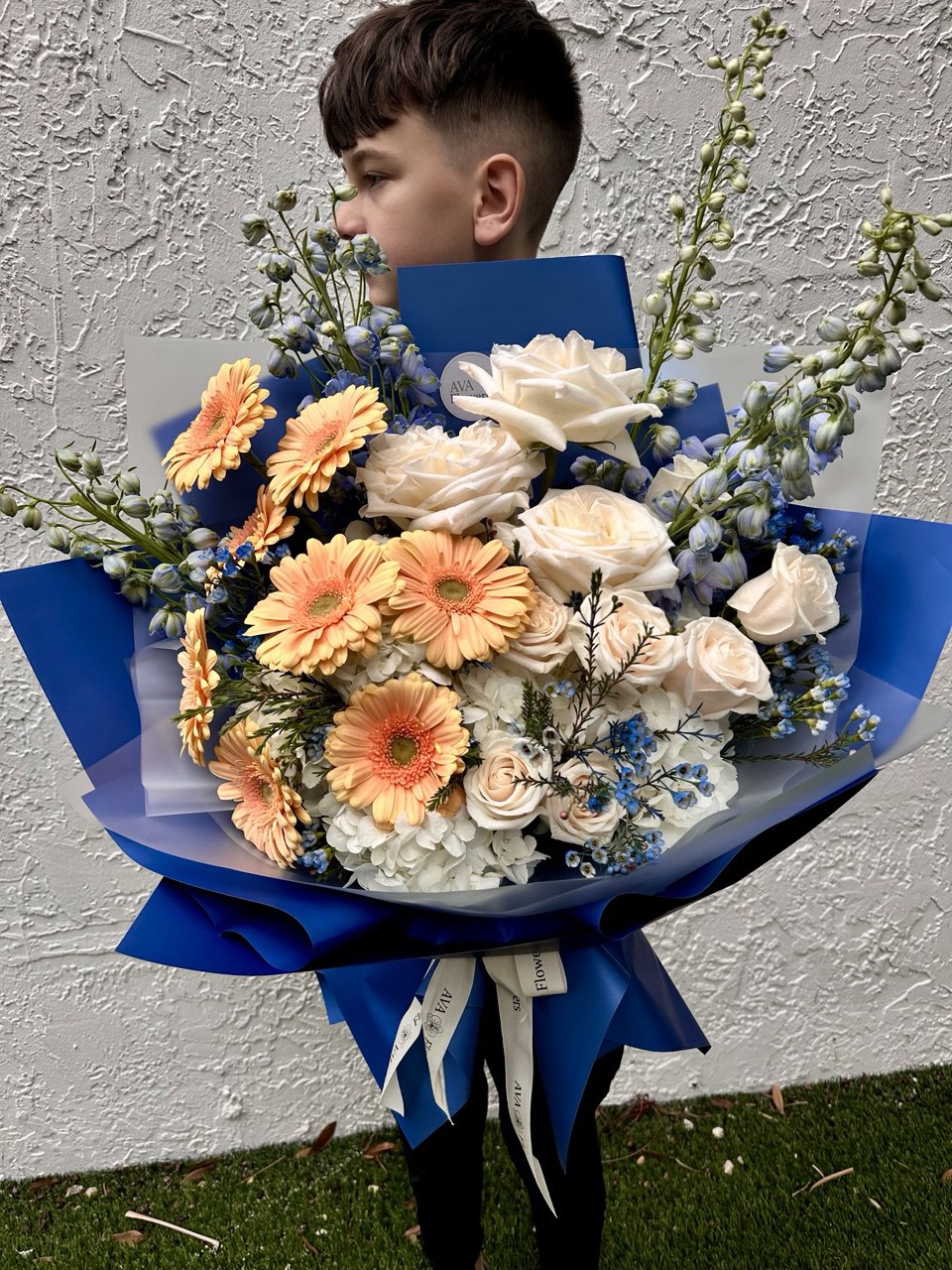Person holding a bouquet of flowers with blue wrapping paper against a light gray wall.