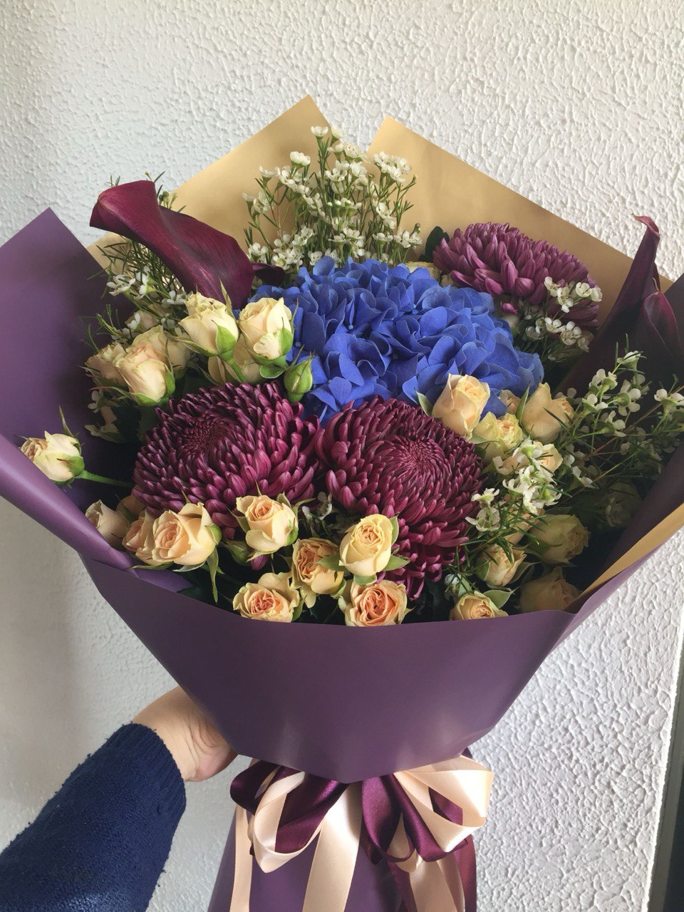 A stylish bouquet featuring deep blue hydrangea, burgundy chrysanthemums, creamy spray roses and white filler flowers, wrapped in purple paper with a satin bow.