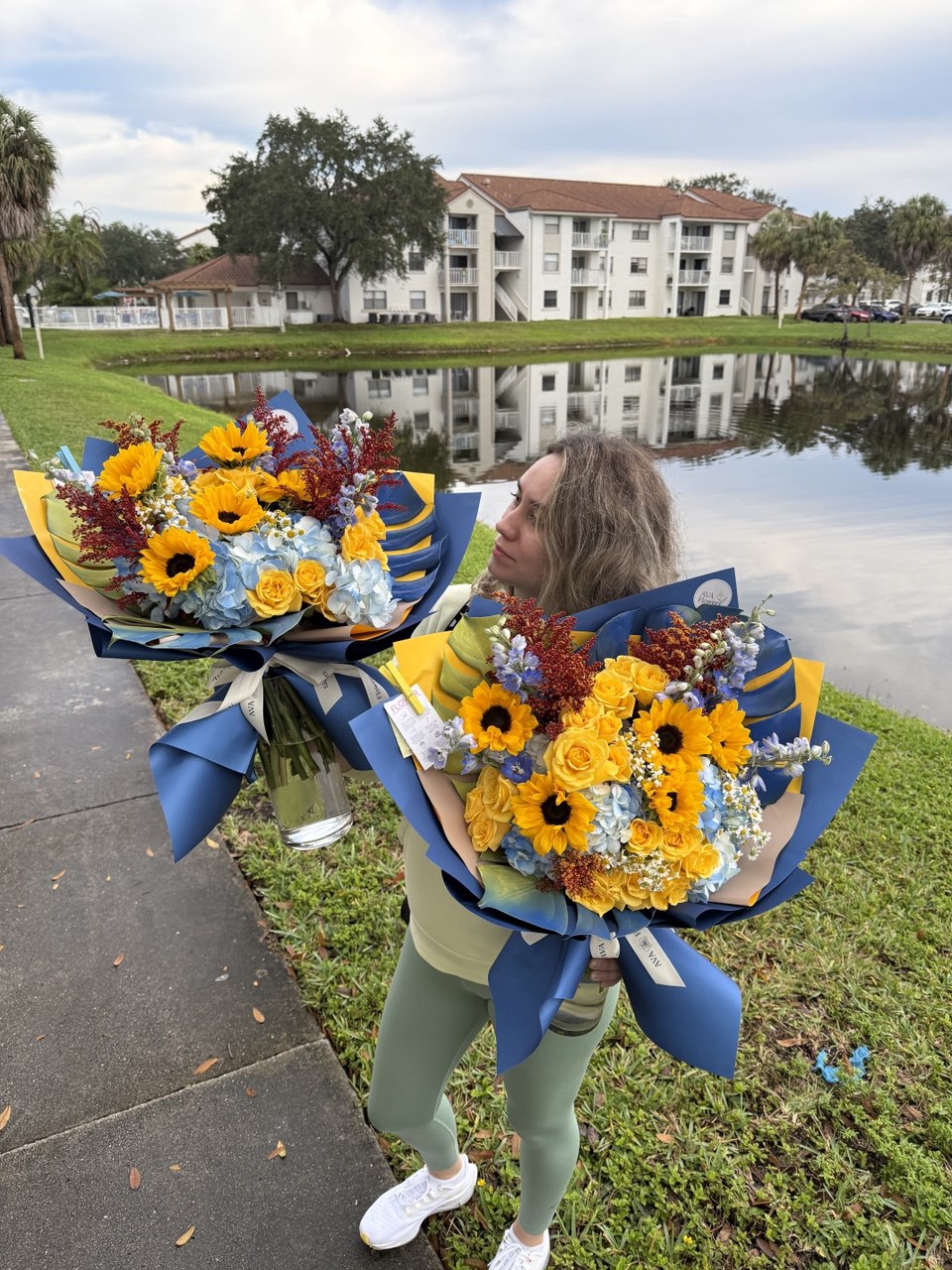 Person holding two large bouquets of flowers by a lake with buildings in the background