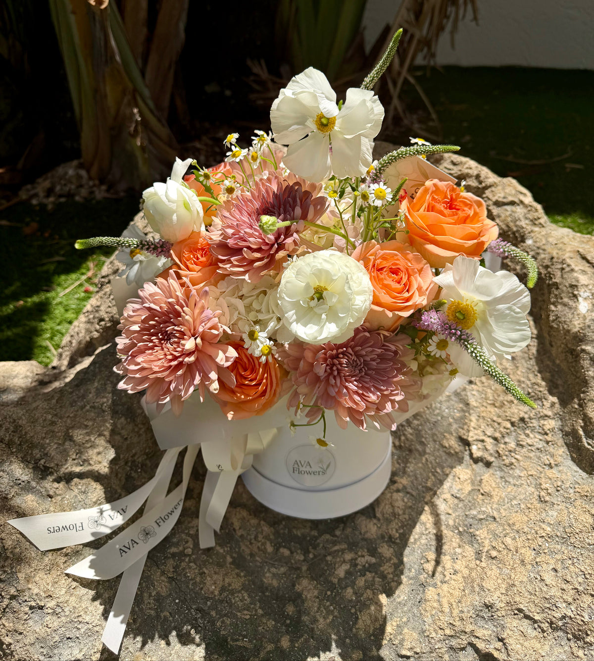 A floral arrangement in a round white box, featuring white, peach, and pink flowers, placed on a large stone outdoors with plants in the background.